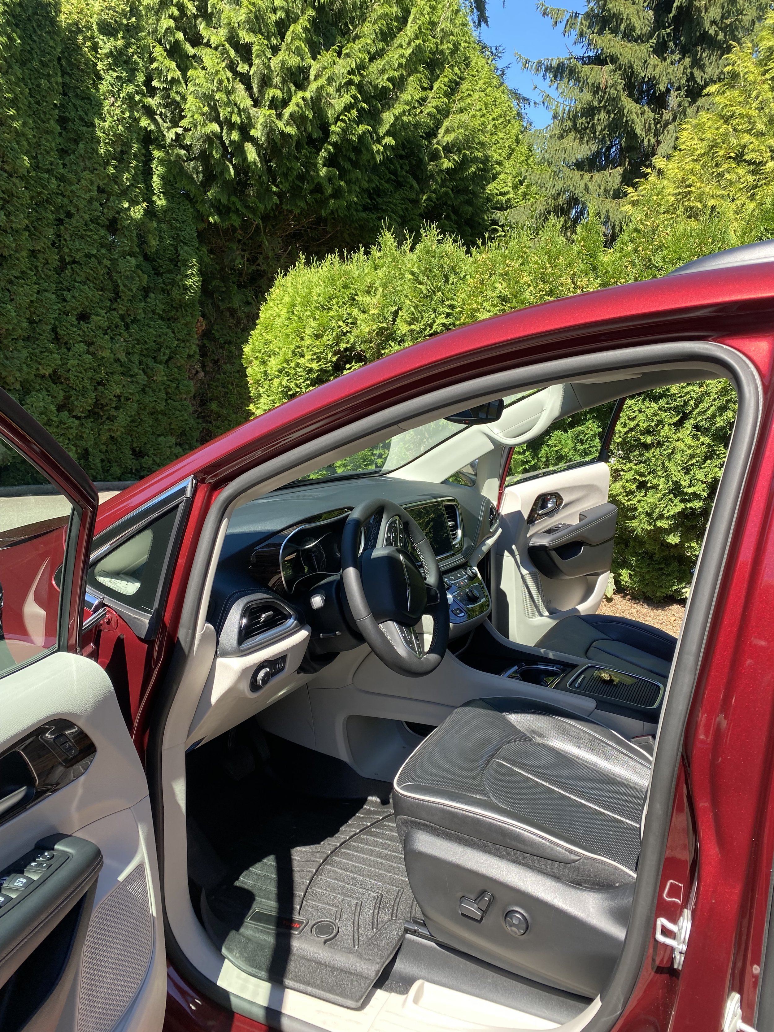 Interior of a red vehicle showing the driver's seat, steering wheel, dashboard, and open door, with green bushes and trees outside on a sunny day.