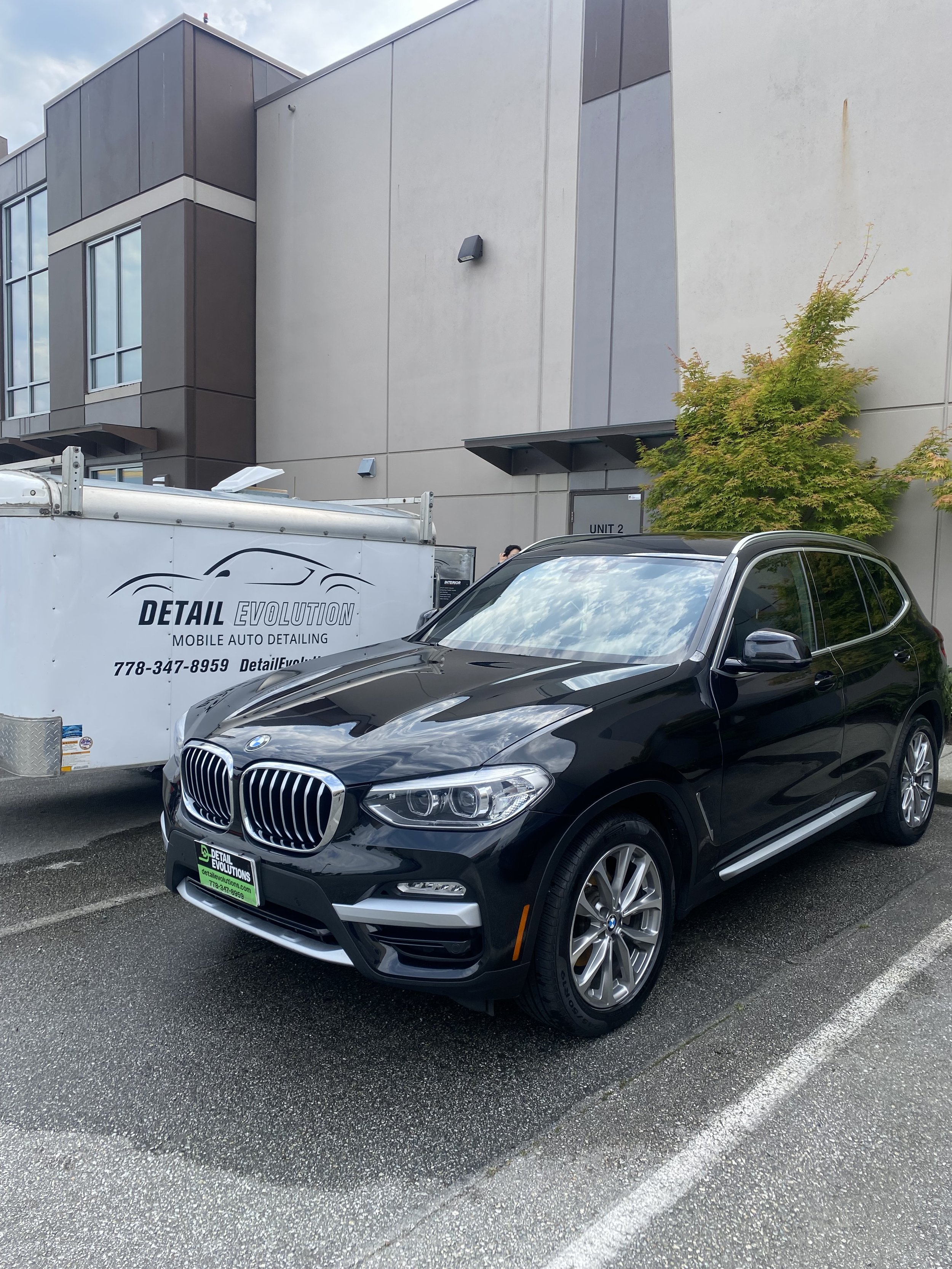 Black BMW SUV parked in a parking lot next to a white trailer with details about an auto detailing service, behind a modern building and a small tree.