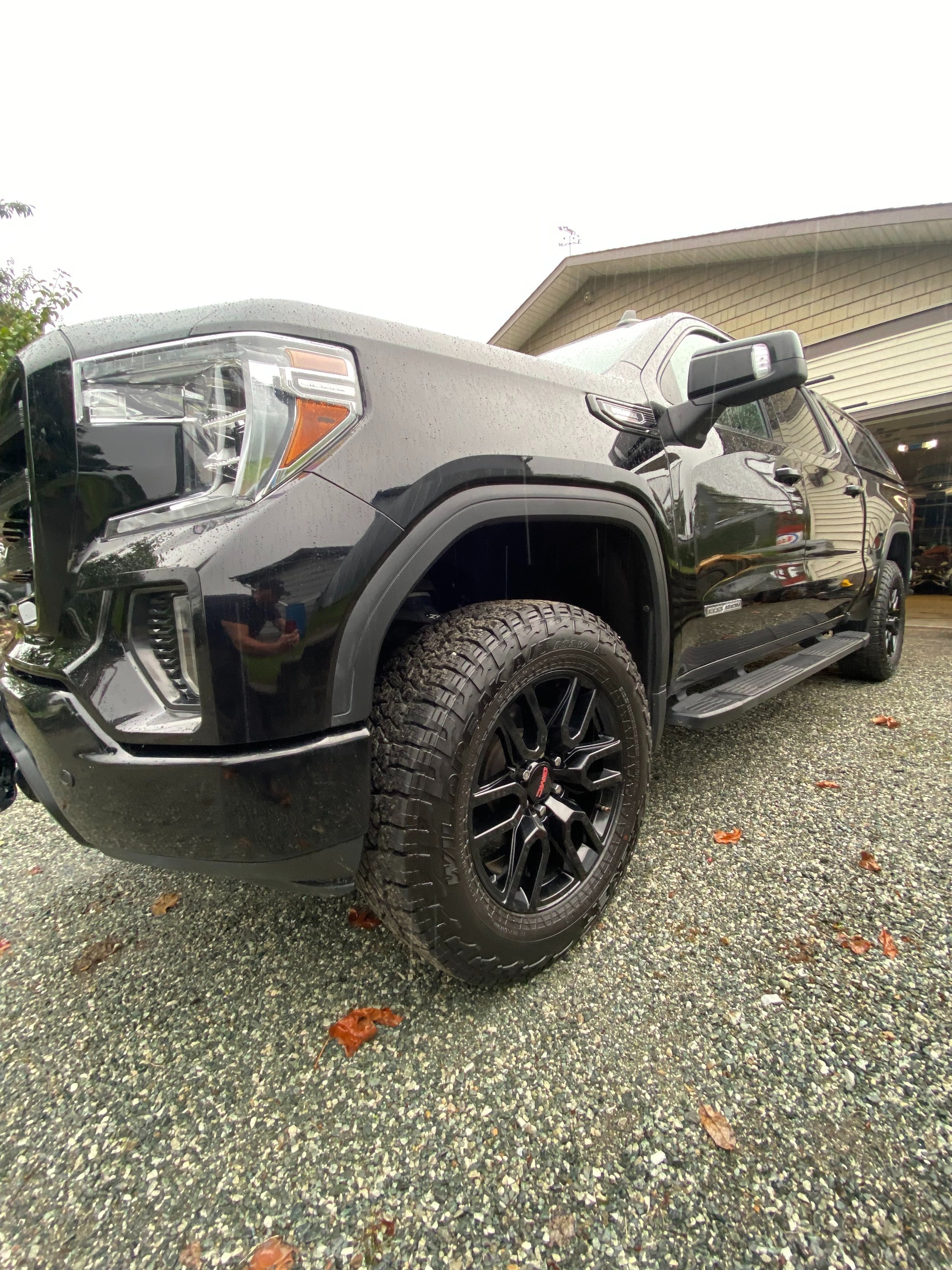Black pickup truck parked in driveway with rain droplets on surface, in front of house at dusk, with wet leaves on gravel ground.