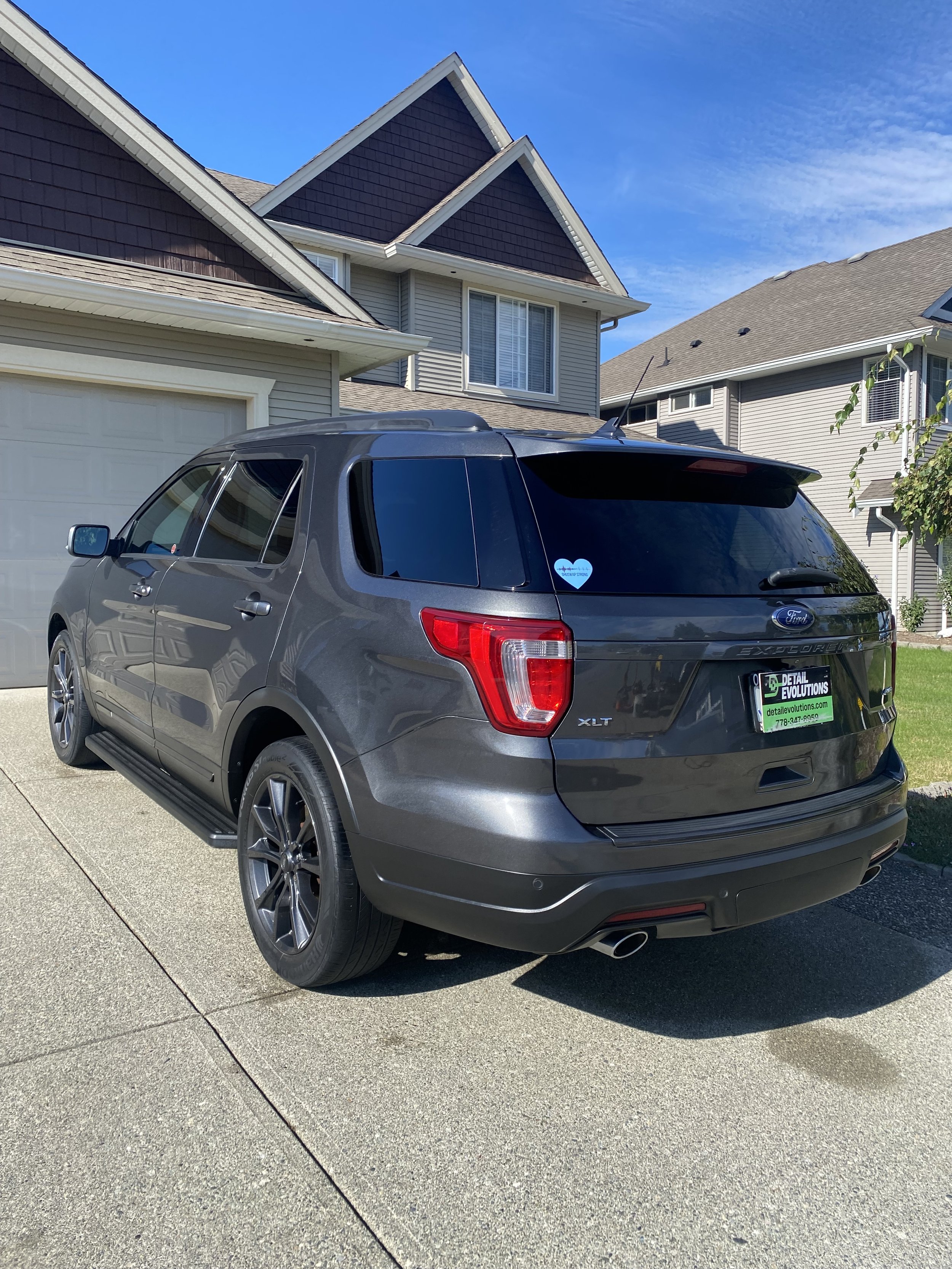 Gray Ford Explorer SUV parked in driveway in front of a multi-story house with beige and brown exterior, under a blue sky.