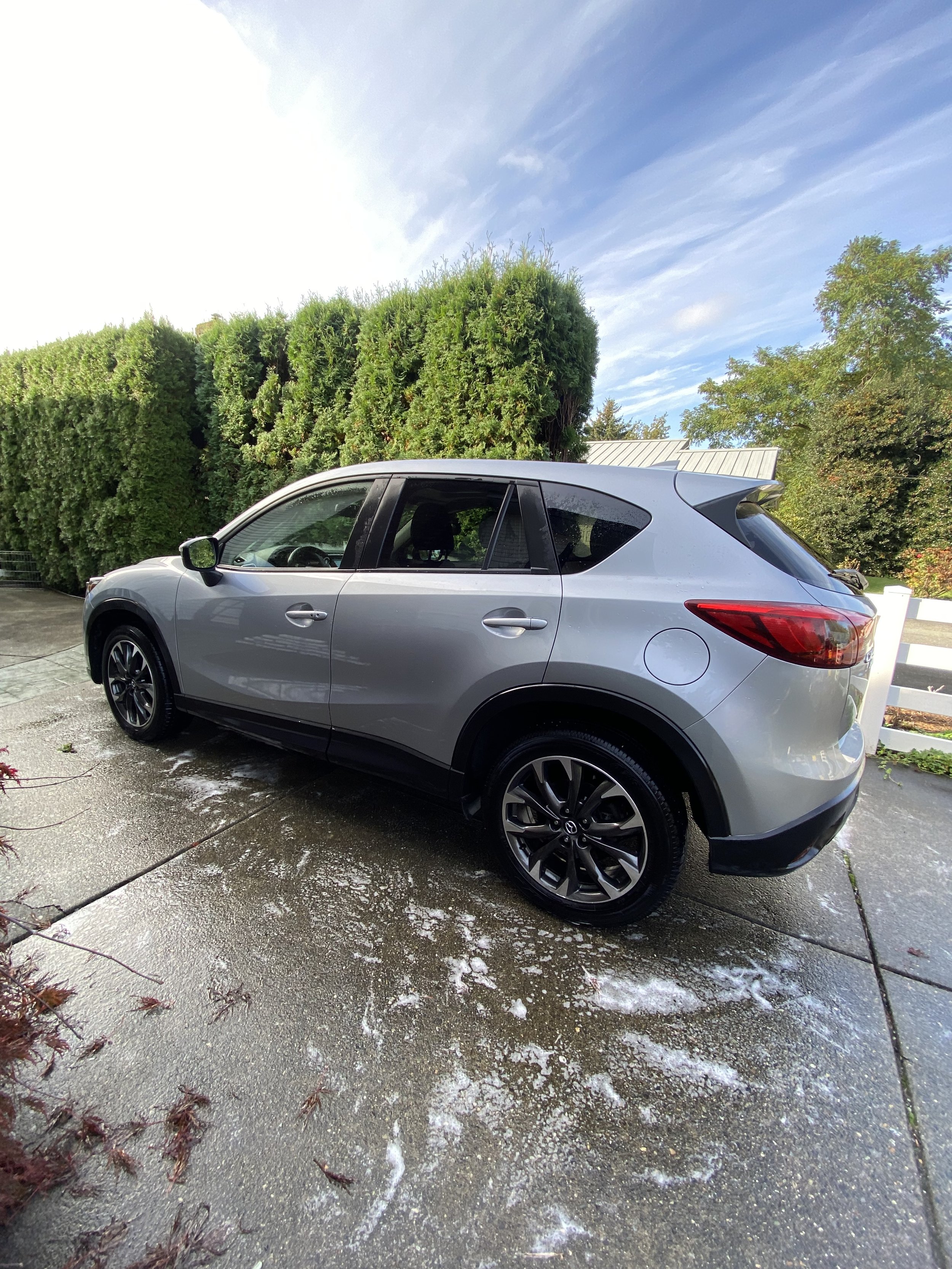 Silver Mazda SUV parked on a wet driveway with soap suds, surrounded by green bushes and trees under a partly cloudy sky.
